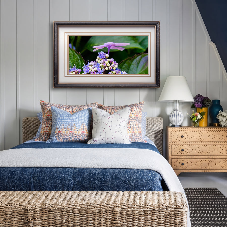 A framed floral art print featuring purple hydrangea flowers and green leaves, displayed on a light gray paneled wall above a neatly made bed with blue and white bedding in a styled bedroom setting.
